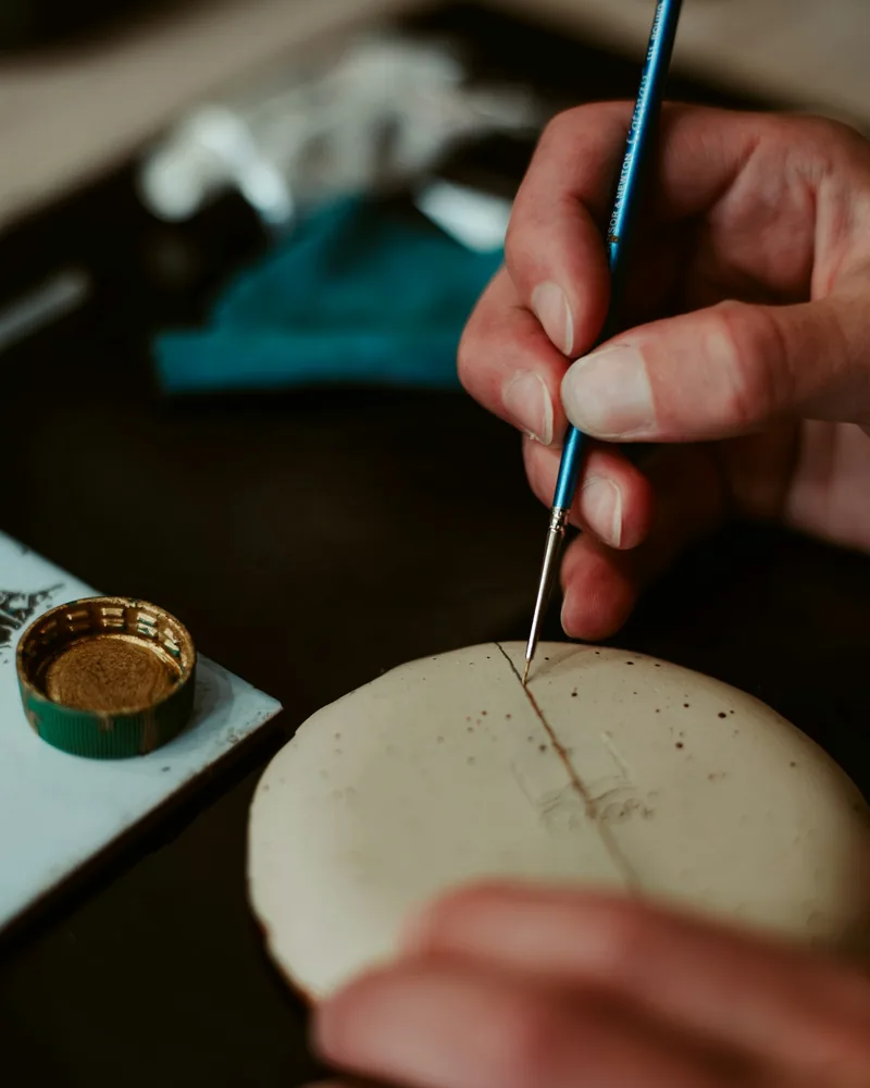 Pincel fino trazando una línea sobre una pieza clara, con un pequeño recipiente de pintura dorada a un lado del banco de trabajo